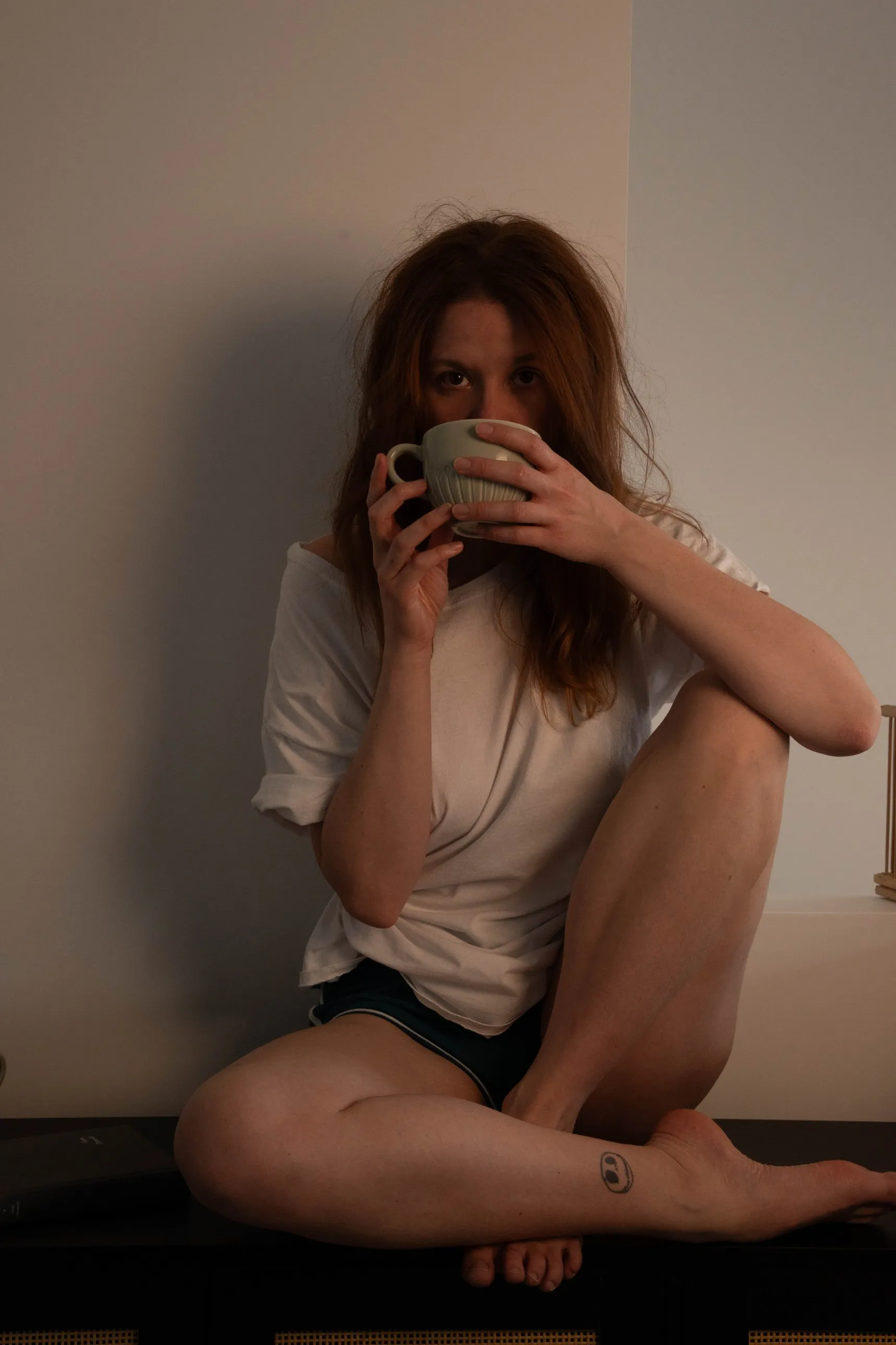 Autoportrait : assise contre un mur, une tasse devant la bouche, le regard droit vers l’objectif
